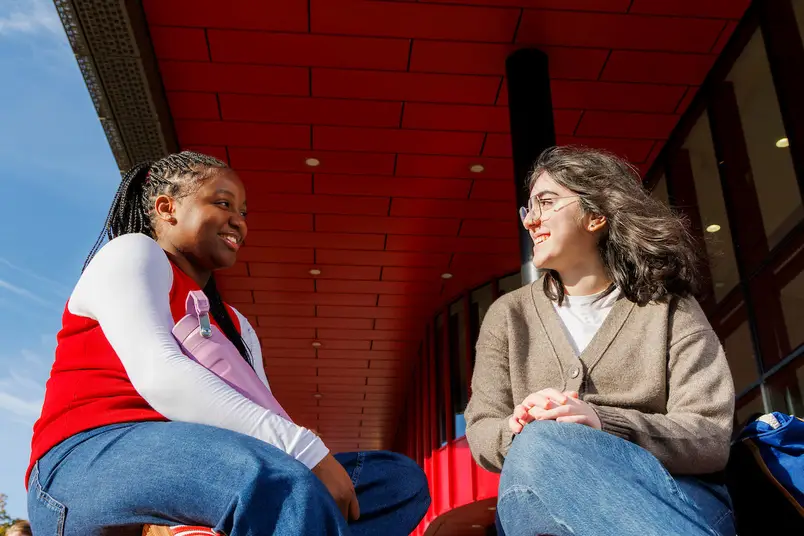 Two students sit facing each other on a bench outside the Eolas building