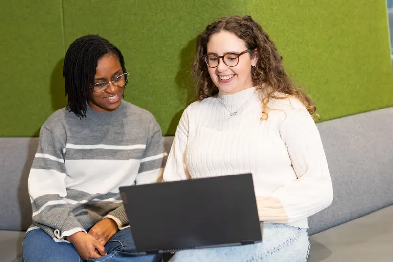 Two students, looking at a laptop, sit in front of a green wall