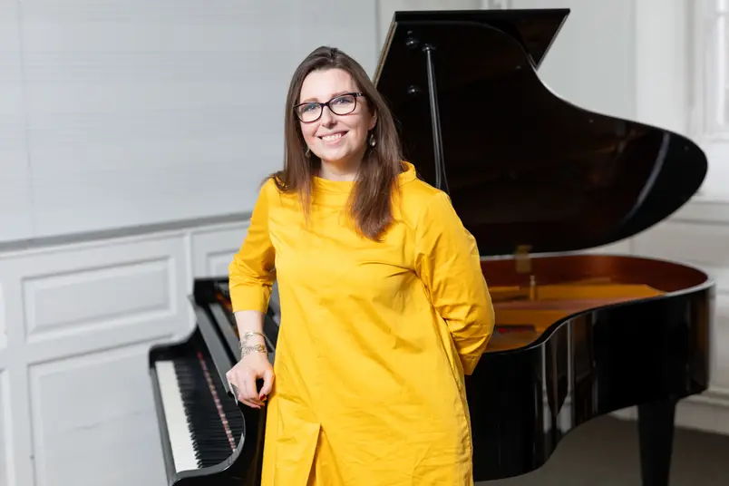 A lecturer in a vibrant yellow dress leans on a grand piano